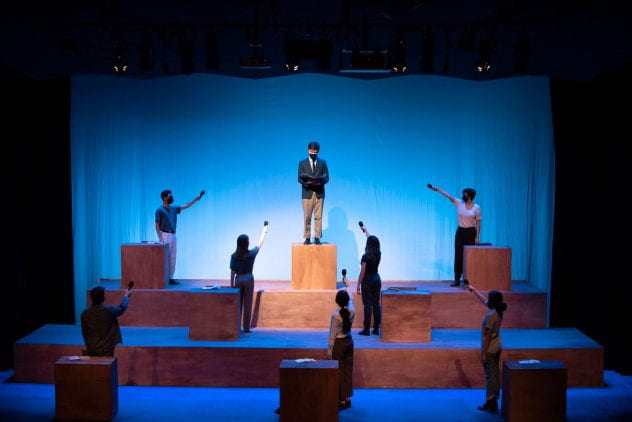 A photo from the Rice Player's production of The Laramie Project. Seven actors point microphones at 1 actor upstage center who stands on a wooden block